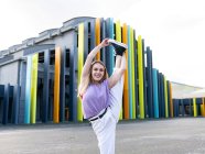 Comprimento total feminino ajuste atraente no desgaste casual realizando de pé dividido e olhando para a câmera com sorriso, enquanto em pé contra a construção criativa no dia ensolarado de verão — Fotografia de Stock