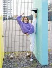 Полная длина положительная гибкая женщина в повседневной одежде выполняя стоя расколоть к бетонной стене и смотреть на камеру, стоя в современном районе — стоковое фото