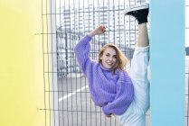 Positive flexible Frau in Freizeitkleidung in voller Länge, die im Stehen gegen eine Betonwand prallt und in die Kamera schaut, während sie in einer modernen Nachbarschaft steht — Stockfoto