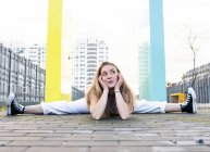Full length flexible pretty female in casual wear doing split and resting head on hand on paved street against creative building on clear summer day — Stock Photo