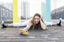 Full body happy fit female doing splits on paved sidewalk and taking selfie on mobile phone in modern urban environment — Stock Photo