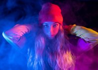 Young confident lady with long blond hair in stylish outfit and hat standing in dark smoky studio with neon illumination and looking at camera — Stock Photo