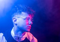 Side view of thoughtful young male in stylish outfit and eyeglasses looking at camera while standing in dark smoky neon room — Stock Photo