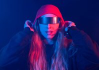 Trendy young female millennial with long blond hair in futuristic sunglasses adjusting hat while standing in dark room with neon illumination — Stock Photo