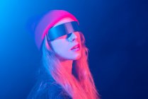 Moda joven milenaria femenina con pelo largo y rubio en gafas de sol futuristas ajustando el sombrero mientras está de pie en habitación oscura con iluminación de neón - foto de stock