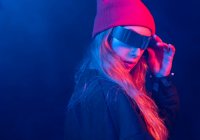 Trendy giovane donna millenaria con lunghi capelli biondi in occhiali da sole futuristici regolazione cappello mentre in piedi in camera buia con illuminazione al neon — Foto stock