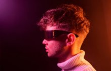 Vista lateral de un hombre joven y seguro de sí mismo con un atuendo elegante que ajusta las gafas de sol futuristas mientras está de pie en un estudio oscuro con iluminación de neón - foto de stock