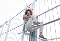 Von unten eine afroamerikanische Athletin in Sportbekleidung, die während des Trainings auf dem Sportplatz auf einer horizontalen Metallstange sitzt und wegschaut — Stockfoto