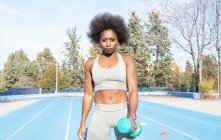 Сосредоточенная черная спортсменка делает упражнения с гантелями во время тренировок на стадионе летом и с нетерпением ждет — стоковое фото