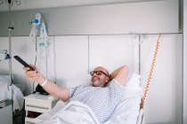 Усміхнений дорослий пацієнт чоловічої статі в лікарняній сукні лежить на ліжку і використовує пульт дистанційного керування в легко обладнаному приміщенні в клініці — стокове фото