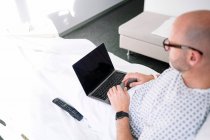 Концентрований дорослий пацієнт чоловічої статі у лікарняній сукні та окулярах, який переглядає нетбук, лежачи на ліжку у світлому залі в клініці — стокове фото