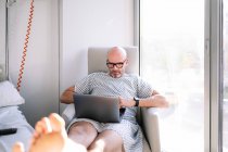 Задумчивый взрослый пациент в больничном платье просматривает современный нетбук, сидя на кресле в солнечном оборудованном отделении клиники — стоковое фото