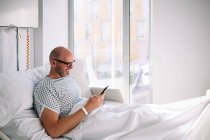 Позитивний дорослий чоловік у сукні для пацієнтів та окулярах, що переглядають телефон на ліжку у світлому залі в сучасній лікарні — стокове фото