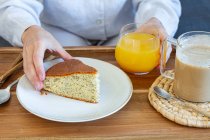 High angle of crop faceless female taking piece of homemade sponge cake from tray with cup of coffee and glass of orange juice during breakfast at home — Stock Photo