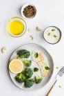 Верхний вид вкусной брокколи со свежим лимоном и соусом возле миски с оливковым маслом и смесью специй на столе — стоковое фото