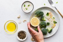 Vista dall'alto di mani di persona irriconoscibili ritagliate che mangiano gustosi broccoli con limone fresco e salsa vicino a ciotole con olio d'oliva e mix di spezie sul tavolo — Foto stock