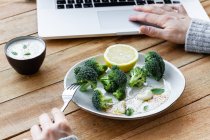 Colheita fêmea anônima com deliciosos brócolos cozidos em garfo navegando na internet no netbook à mesa — Fotografia de Stock