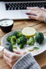 Crop anonymous female with delicious cooked broccoli on fork browsing internet on netbook at table — Stock Photo