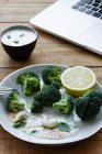 Deliziosi broccoli cotti con fetta di limone e anacardi vicino alla ciotola con salsa bianca e netbook su tavolo di legno — Foto stock