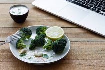Delicioso brócoli cocido con rodaja de limón y nueces de anacardo cerca de un tazón con salsa blanca y netbook en la mesa de madera - foto de stock