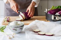 Неузнаваемый шеф-повар в фартуке режет баклажаны ножом на разделочной доске во время приготовления здорового обеда на кухне — стоковое фото