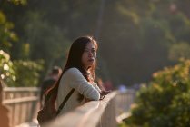 На схилі видніється дбайлива молода азіатка, одягнена в повсякденний одяг, що спирається на поруччя пішохідного мосту і озирається в буйних лісах в сонячний день. — стокове фото