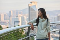 Обнаженная молодая женщина азиатского происхождения в повседневной одежде стоит у перил и отводит взгляд в светлое время суток в современном городском городе — стоковое фото