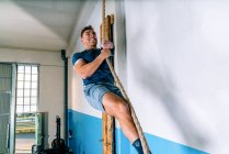 Side view of disabled male athlete in sports clothes climbing workout rope near bright wall in gym — Stock Photo