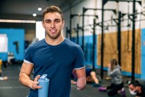 Young disabled happy male athlete in sportswear holding bottle of water while looking at camera during workout near unrecognizable partners in gymnasium — Stock Photo