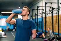 Young disabled male athlete in sportswear drinking beverage from bottle and looking up during workout near unrecognizable partners in gymnasium — Stock Photo