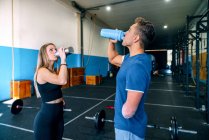 Side view of young sportswoman near handicapped male athlete drinking water from bottles while looking up during workout near barbells in gym — Stock Photo