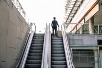 Low angle back view of anonymous male traveler with suitcase standing on moving stairs near building of airport — Stock Photo