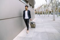 Grave viaggiatore nero maschio in abito alla moda e maschera a piedi con valigia in città e guardando altrove — Foto stock