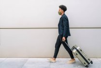 Вид збоку чорний чоловічий мандрівник у модному вбранні та масці, що йде з валізою у місті та дивиться в сторону — стокове фото