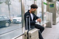 Vista lateral de viajando afro-americano macho em máscara protetora sentado no banco com mala e verificação da hora de chegada do trem — Fotografia de Stock