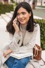 Fröhliche Unternehmerin sitzt auf Bank in der Stadt und telefoniert, während sie lächelt und wegschaut — Stockfoto