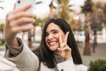 Sonriente encantadora hembra tomando selfie en el teléfono inteligente mientras está de pie en la calle con palmeras - foto de stock