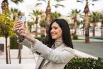 Sonriente encantadora hembra tomando selfie en el teléfono inteligente mientras está de pie en la calle con palmeras - foto de stock