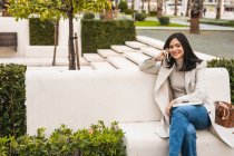 Весела жінка-підприємець сидить на лавці в місті і розмовляє на мобільному телефоні, посміхаючись і дивлячись — стокове фото
