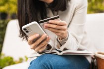 Recortar hembra sentado en el banco y hacer la compra con tarjeta de plástico a través de teléfono inteligente durante las compras en línea - foto de stock