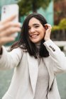 Smiling charming female taking selfie on smartphone while standing in street with palm trees — Stock Photo