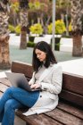 Femme entrepreneure sérieuse assise sur un banc dans un parc urbain et tapant sur un ordinateur portable tout en travaillant à distance sur un projet d'entreprise — Photo de stock