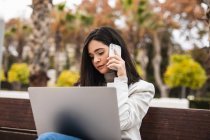 Концентрована бізнес-леді сидить на лавці з ноутбуком і розмовляє смартфоном під час віддаленої роботи в міському парку — стокове фото