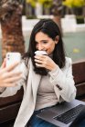 Entzückte junge Unternehmerin sitzt mit Laptop auf Bank und macht Selbstporträt mit Coffee to go in Pappbecher, während sie das Smartphone benutzt — Stockfoto