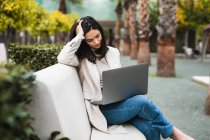 Entrepreneur féminin pensif utilisant un ordinateur portable tout en étant assis sur le banc dans le parc de la ville et de travailler en ligne sur le projet — Photo de stock
