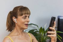 Allegro femmina adulta in auricolari che hanno videochiamata attraverso il moderno telefono cellulare in soggiorno leggero — Foto stock
