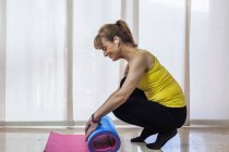 Side view full length smiling adult female in sportswear listening to favorite music and rolling yoga mat after intense training in light fitness center — Stock Photo