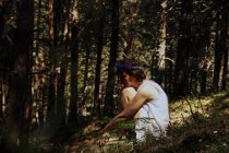 Вид збоку романтичної молодої леді з коротким волоссям в літньому платті і квітковим вінком, що приймає коліна, сидячи на траві в пишному лісі — стокове фото