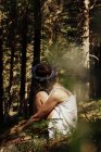 Seitenansicht von unkenntlich romantische nachdenkliche Dame mit kurzen Haaren im Sommerkleid und Blumenkranz umarmen Knie, während sie auf Gras im üppigen Wald sitzen — Stockfoto