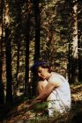 Vue latérale de romantique réfléchie jeune femme aux cheveux courts et les yeux fermés en robe d'été et couronne florale embrassant les genoux tout en étant assis sur l'herbe dans la forêt luxuriante — Photo de stock